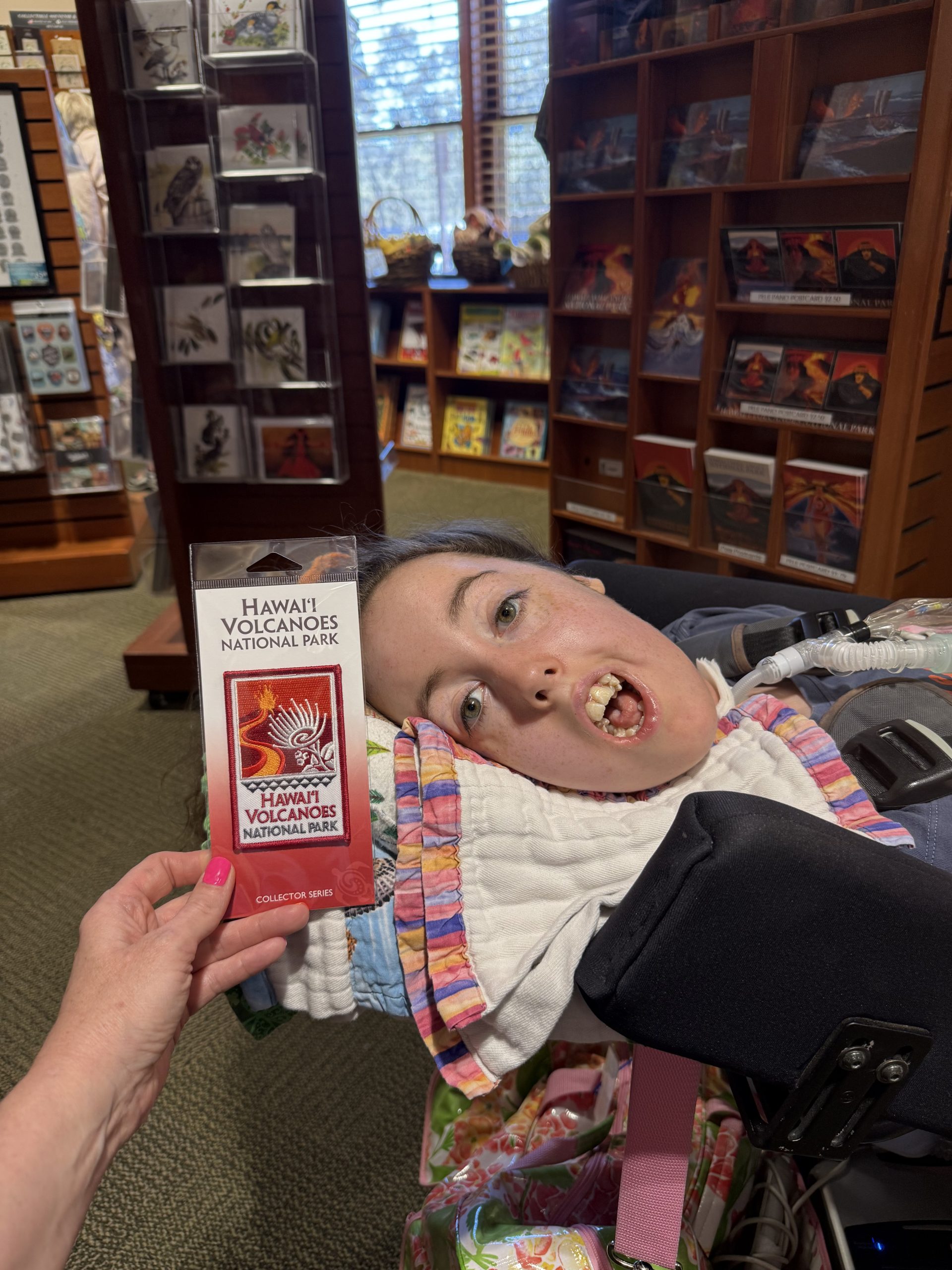 A little girl in a wheelchair in a bookstore with a hand holding up a patch in front of her that reads, "Hawai'i Volcanoes National Park."