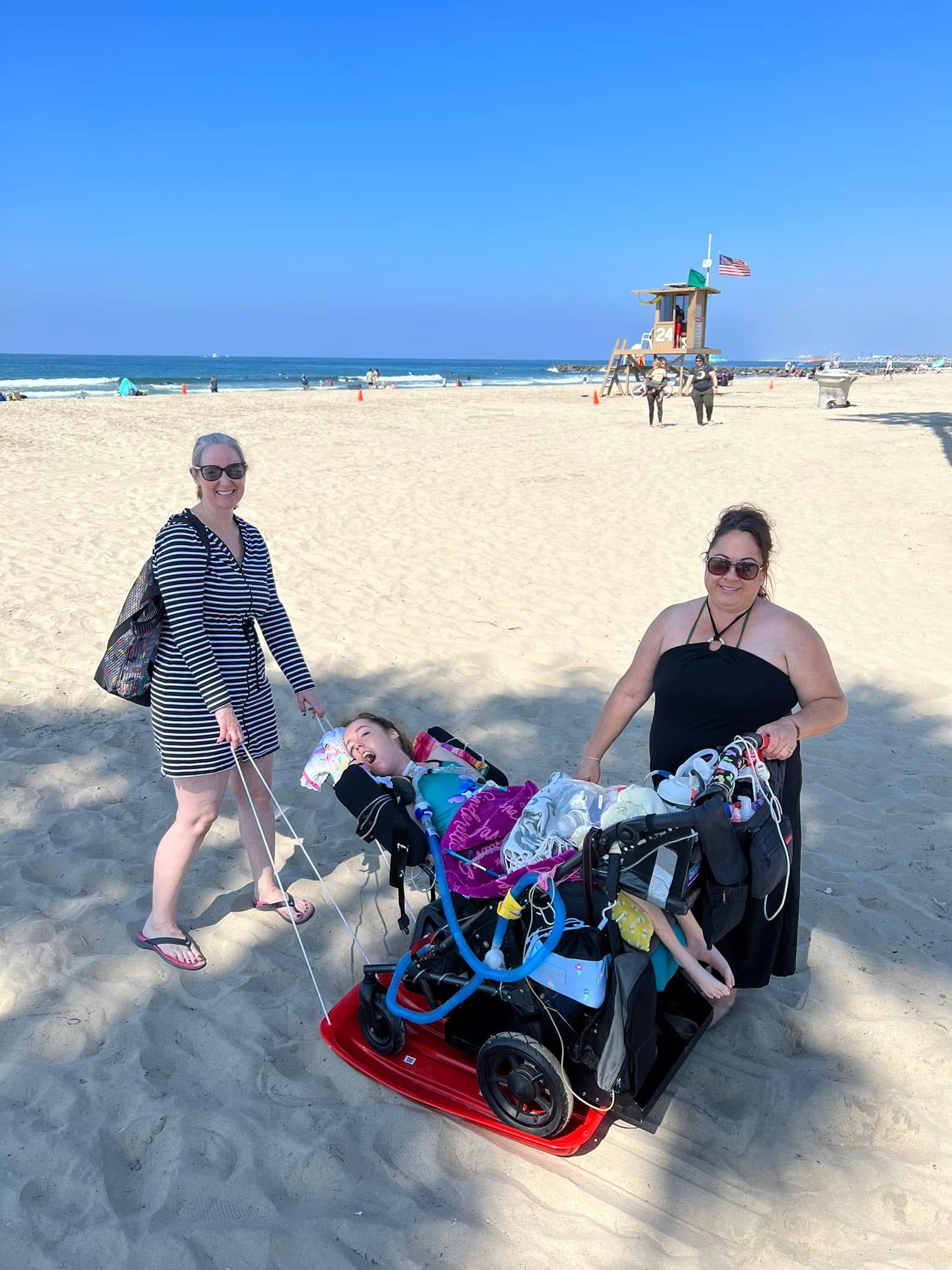 Kennedy, Jennifer, and a friend at the beach. Jennifer is pulling Kennedy's wheelchair with a sled over the sand.
