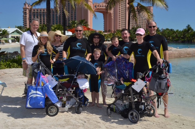 A large, happy group at a beach resort. Everybody is in "Dolphic Cay" wetsuits, and two children are in wheelchairs.