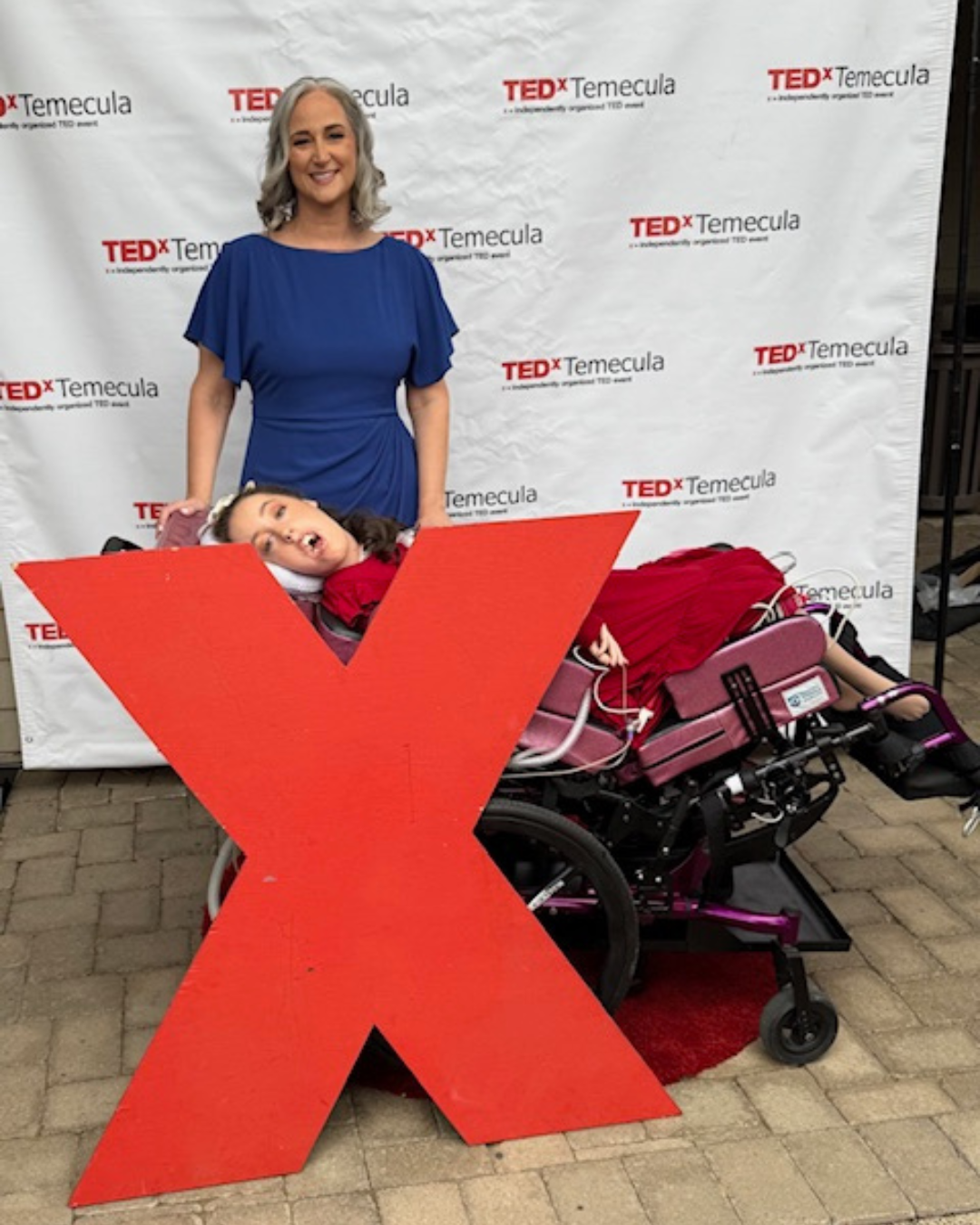 Jennifer in a blue dress standing behind a Kennedy in a purple wheelchair, in front of a TEDx Temecula backdrop and large red X.
