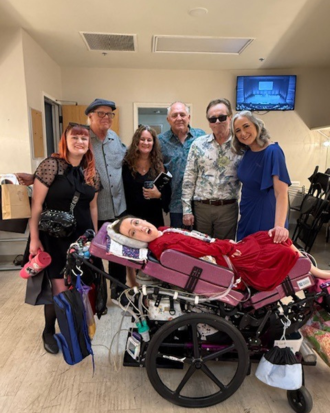 Group photo indoors with six adults standing behind Kennedy reclined in a purple wheelchair wearing a red dress.