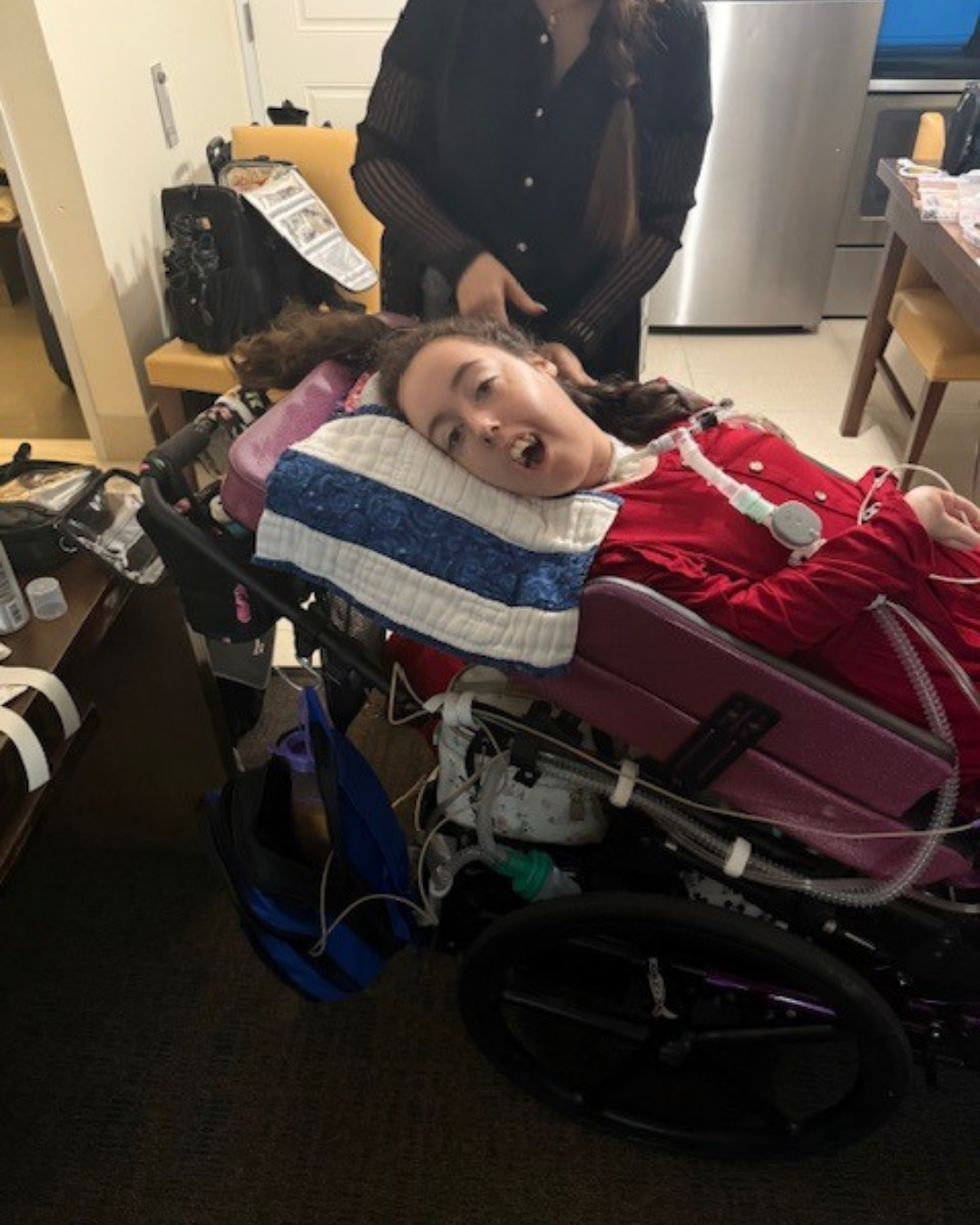 Caregiver braiding a Kennedy's hair while she rests in a reclining purple wheelchair in a kitchen/dining area.