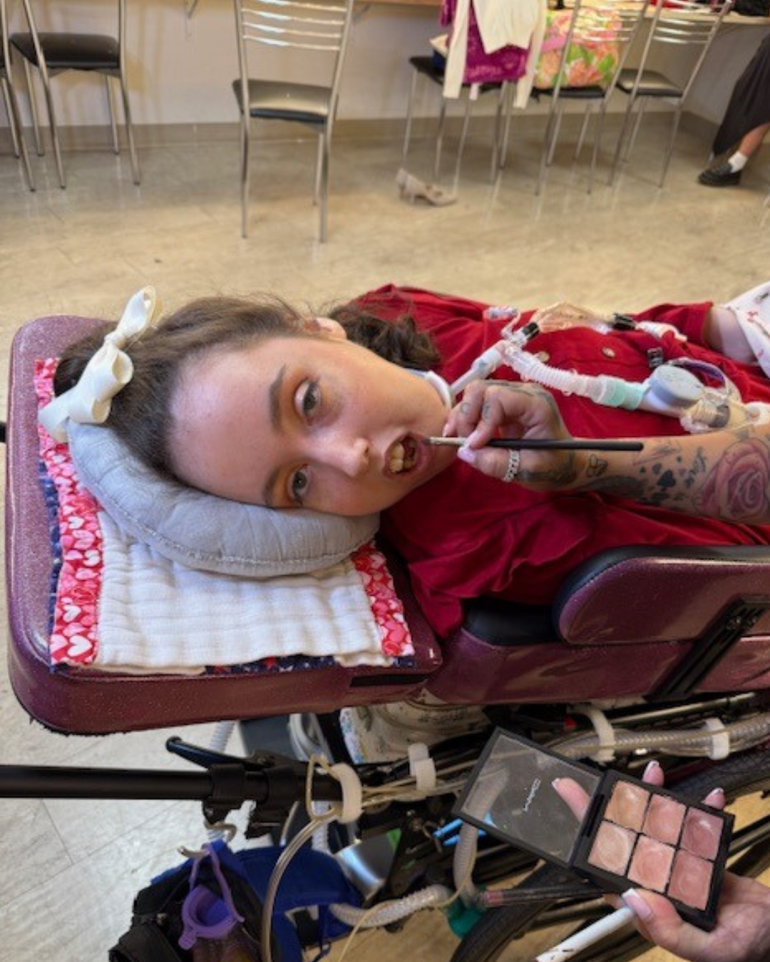 Kennedy reclined in a purple wheelchair having lipstick applied with a brush; makeup palette visible in the foreground.
