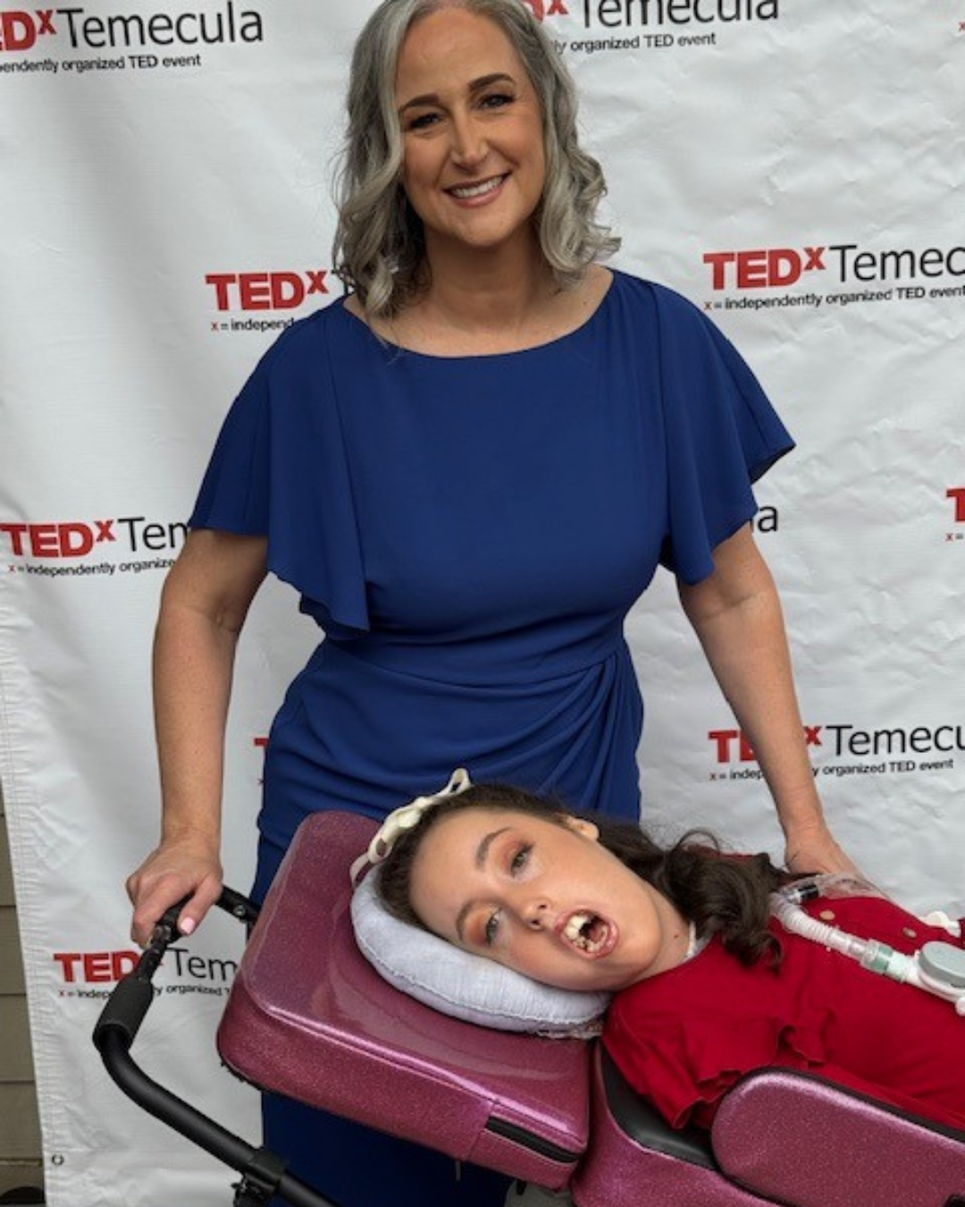 Jennifer, in a blue dress, standing over Kennedy in a purple wheelchair at the TEDx conference.