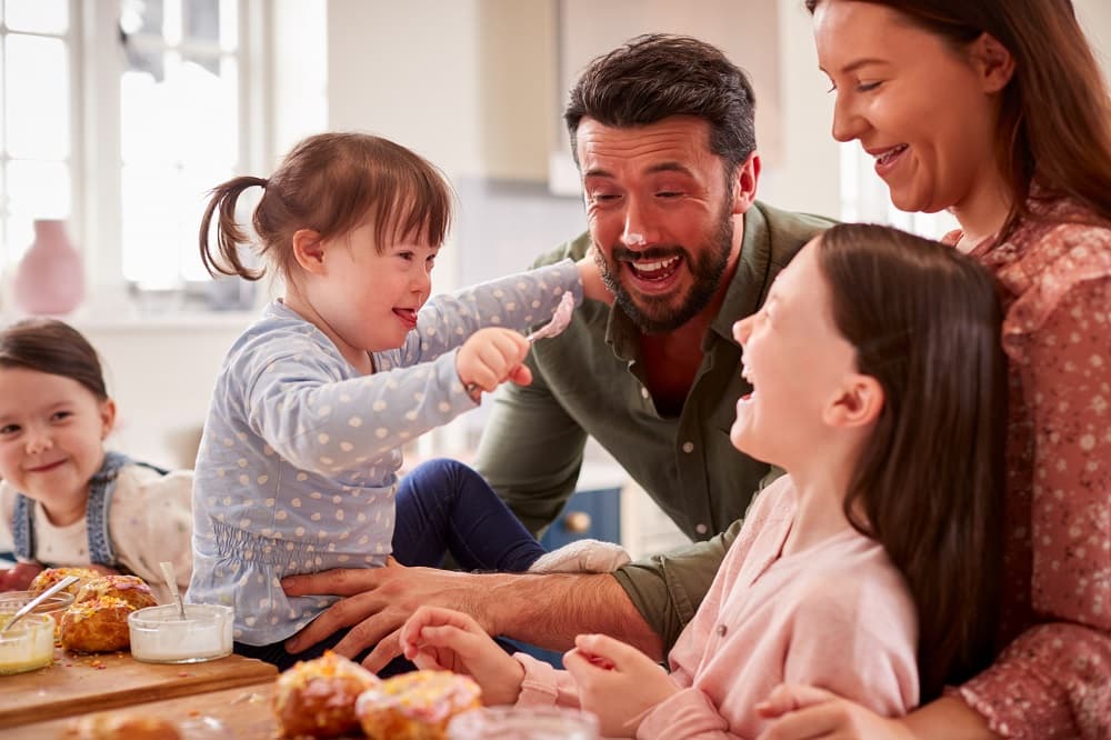 Parents and children laughing and interacting around a table with food in a bright kitchen.