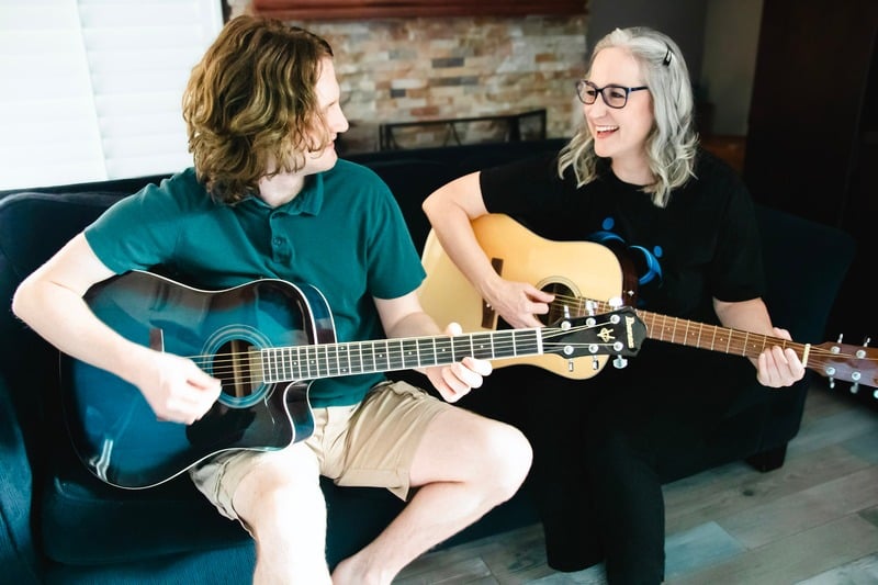 A woman and a young man sitting on a couch, playing acoustic guitars together and smiling in a relaxed home setting.
