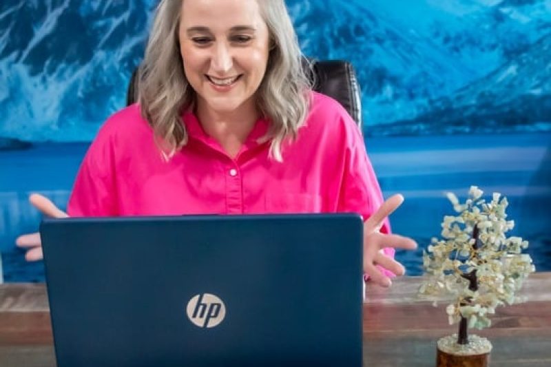 A smiling woman in a pink shirt working on a laptop at a desk, with a scenic mountain and lake backdrop behind her.